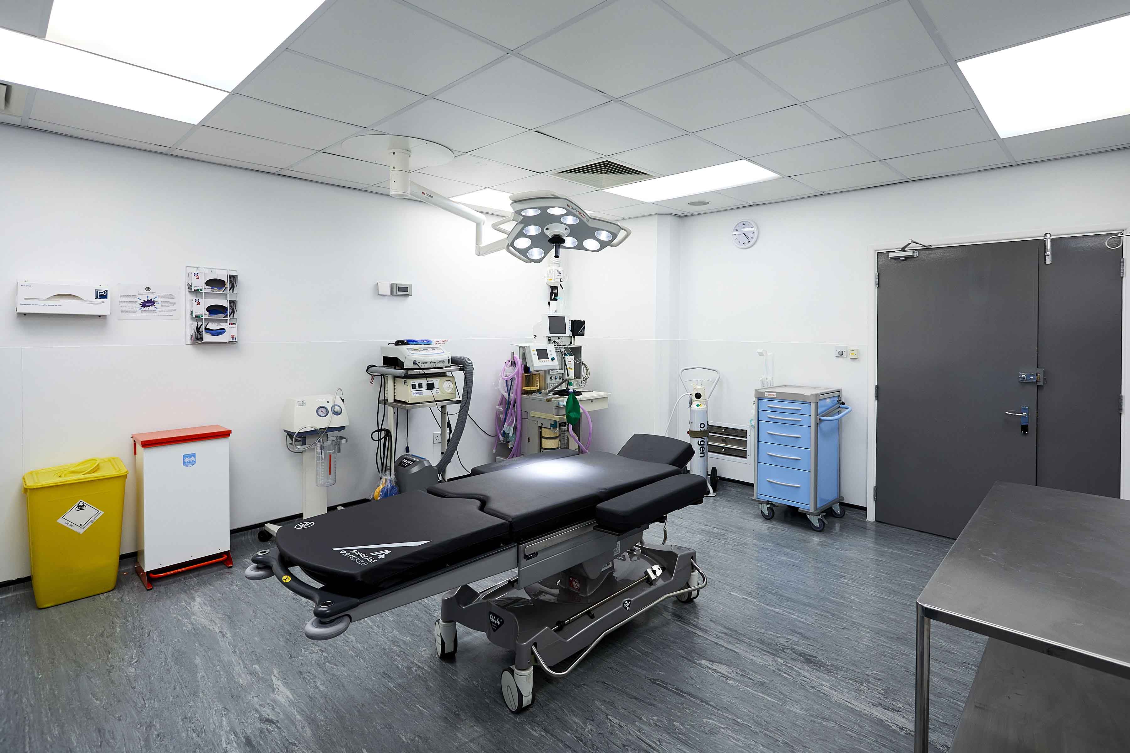 A clean surgical theatre with a black operating table, overhead surgical light, medical equipment trolleys, and a yellow clinical waste bin.