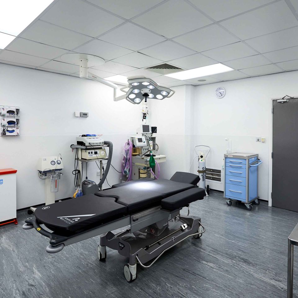 A clean surgical treatment room with a black operating table, overhead surgical light, medical equipment trolleys, and an oxygen cylinder.