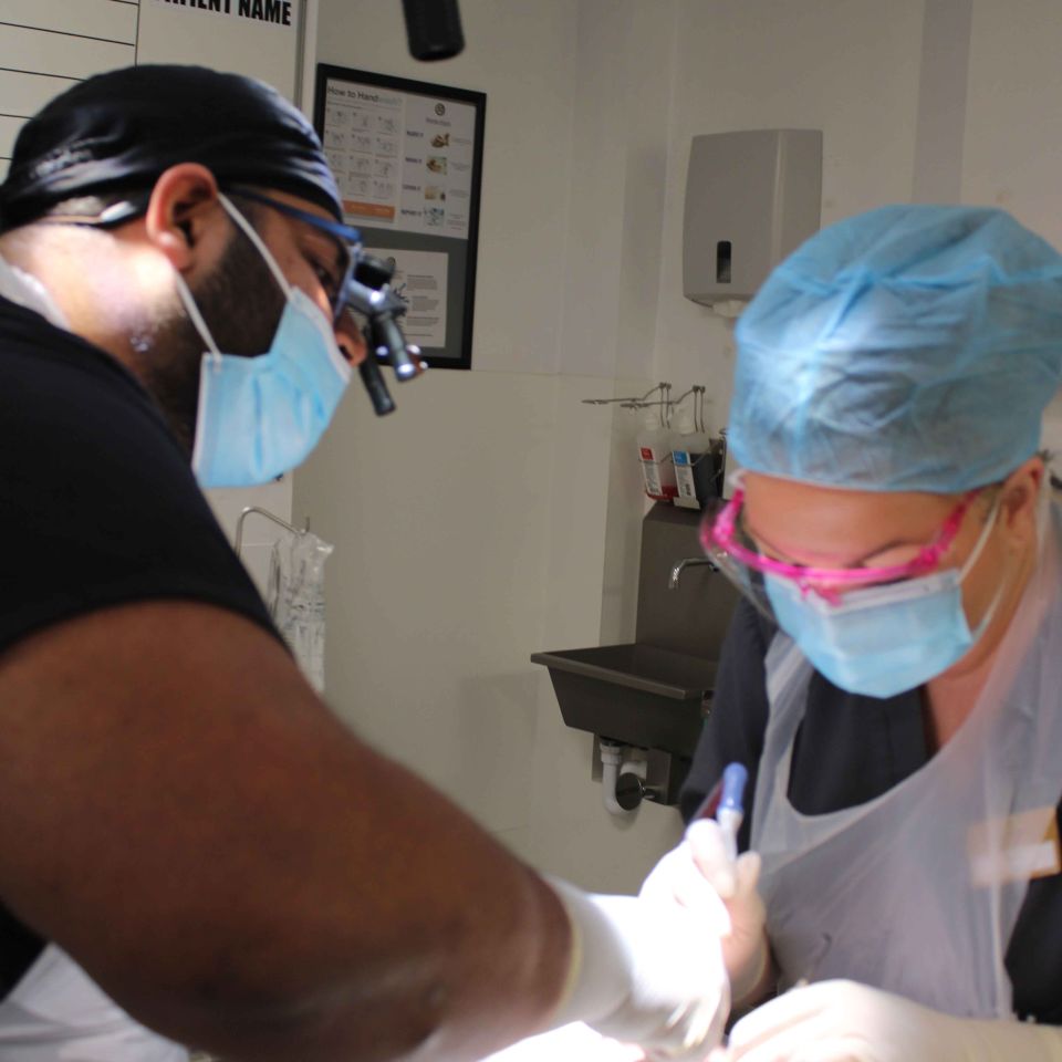 Two dental surgeons in masks and surgical caps performing a procedure on a patient under theatre lights