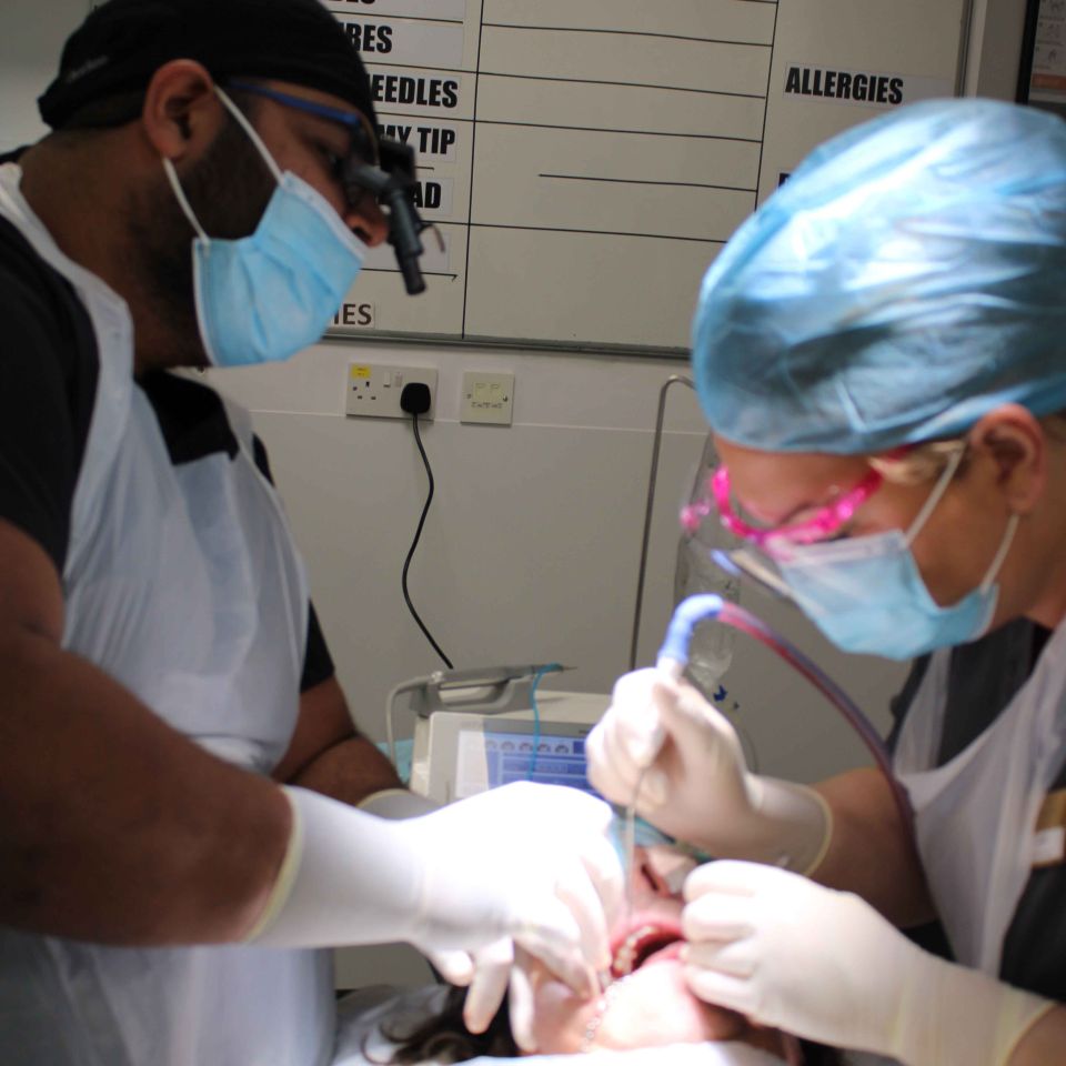Two masked dental surgeons in gloves and scrubs performing a tooth extraction procedure on a patient in a clinical setting.