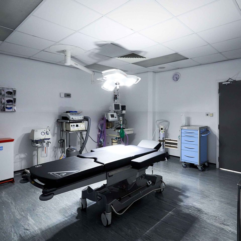 Empty surgical operating theatre with a black patient table, overhead surgical light, anaesthesia equipment, and medical trolley